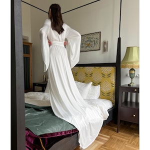 Op de afbeelding: Een witte satijnen badjas met lange mouwen en een strikceintuur. De badjas is gedrapeerd over een bed met een hoofdeinde met bloemenpatroon.