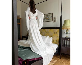 Può includere: Una vestaglia di raso bianco con maniche lunghe e una cintura a nodo. La vestaglia è drappeggiata su un letto con una testiera a motivi floreali.