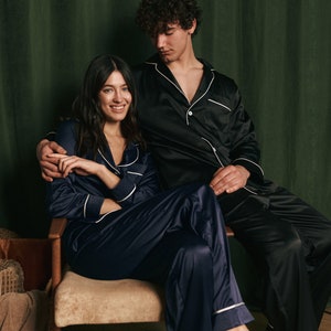 K&ouml;nnte beinhalten: Ein Paar tr&auml;gt passende Satin-Pyjamas. Die Frau sitzt auf einem Stuhl mit gekreuzten Beinen, w&auml;hrend der Mann hinter ihr steht und seinen Arm um ihre Taille legt. Beide l&auml;cheln und schauen in die Kamera.