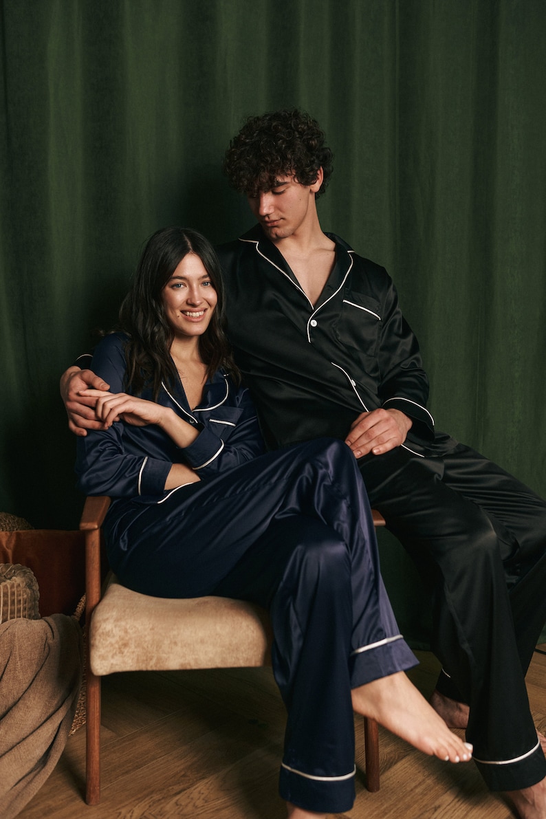 K&ouml;nnte beinhalten: Ein Paar tr&auml;gt passende Satin-Pyjamas. Die Frau sitzt auf einem Stuhl mit gekreuzten Beinen, w&auml;hrend der Mann hinter ihr steht und seinen Arm um ihre Taille legt. Beide l&auml;cheln und schauen in die Kamera.