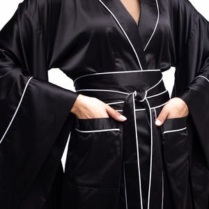 May include: A black satin robe with white piping. The robe has a shawl collar, long sleeves, and a tie belt. The robe has pockets on the front.