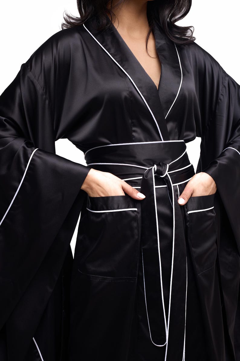 May include: A black satin robe with white piping. The robe has a shawl collar, long sleeves, and a tie belt. The robe has pockets on the front.