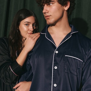 K&ouml;nnte beinhalten: Ein Paar tr&auml;gt passende Satin-Pyjamas. Der Mann tr&auml;gt ein marineblaues Pyjama-Oberteil mit wei&szlig;en Paspeln und die Frau tr&auml;gt ein schwarzes Pyjama-Oberteil mit rosa Paspeln.