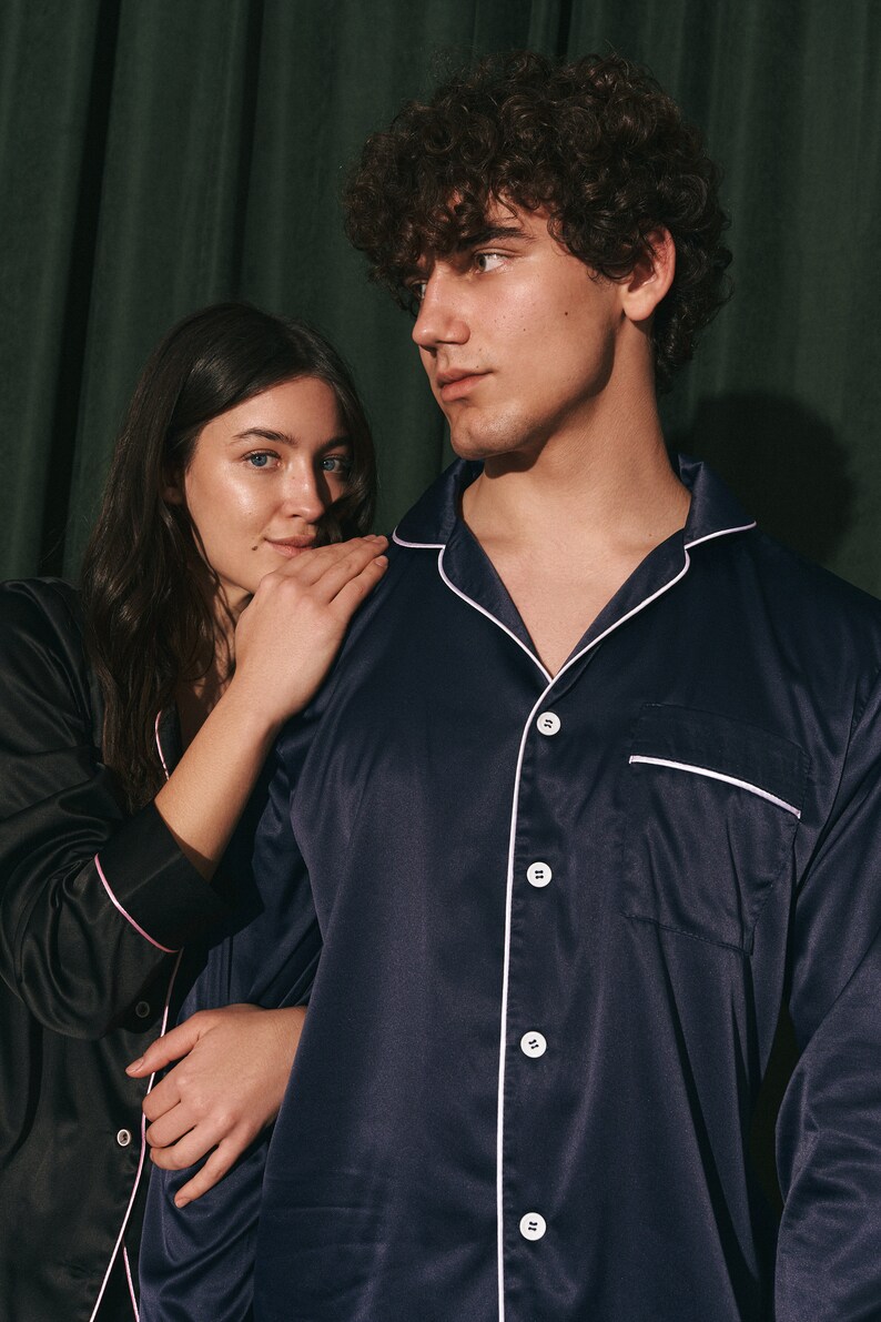 K&ouml;nnte beinhalten: Ein Paar tr&auml;gt passende Satin-Pyjamas. Der Mann tr&auml;gt ein marineblaues Pyjama-Oberteil mit wei&szlig;en Paspeln und die Frau tr&auml;gt ein schwarzes Pyjama-Oberteil mit rosa Paspeln.