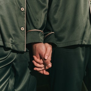 K&ouml;nnte beinhalten: Zwei Personen tragen passende dunkelgr&uuml;ne Satin-Pyjamas mit wei&szlig;en Paspeln. Sie halten sich an den H&auml;nden.