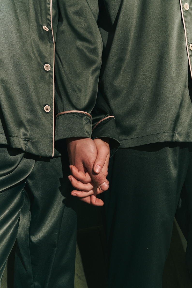 K&ouml;nnte beinhalten: Zwei Personen tragen passende dunkelgr&uuml;ne Satin-Pyjamas mit wei&szlig;en Paspeln. Sie halten sich an den H&auml;nden.