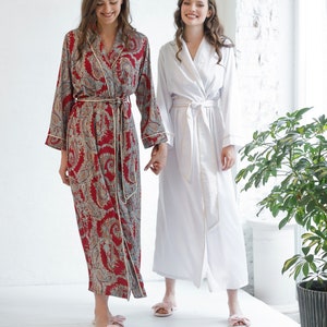 May include: Two women wearing long robes, one in a white robe and the other in a burgundy paisley robe. Both women are wearing pink slippers.