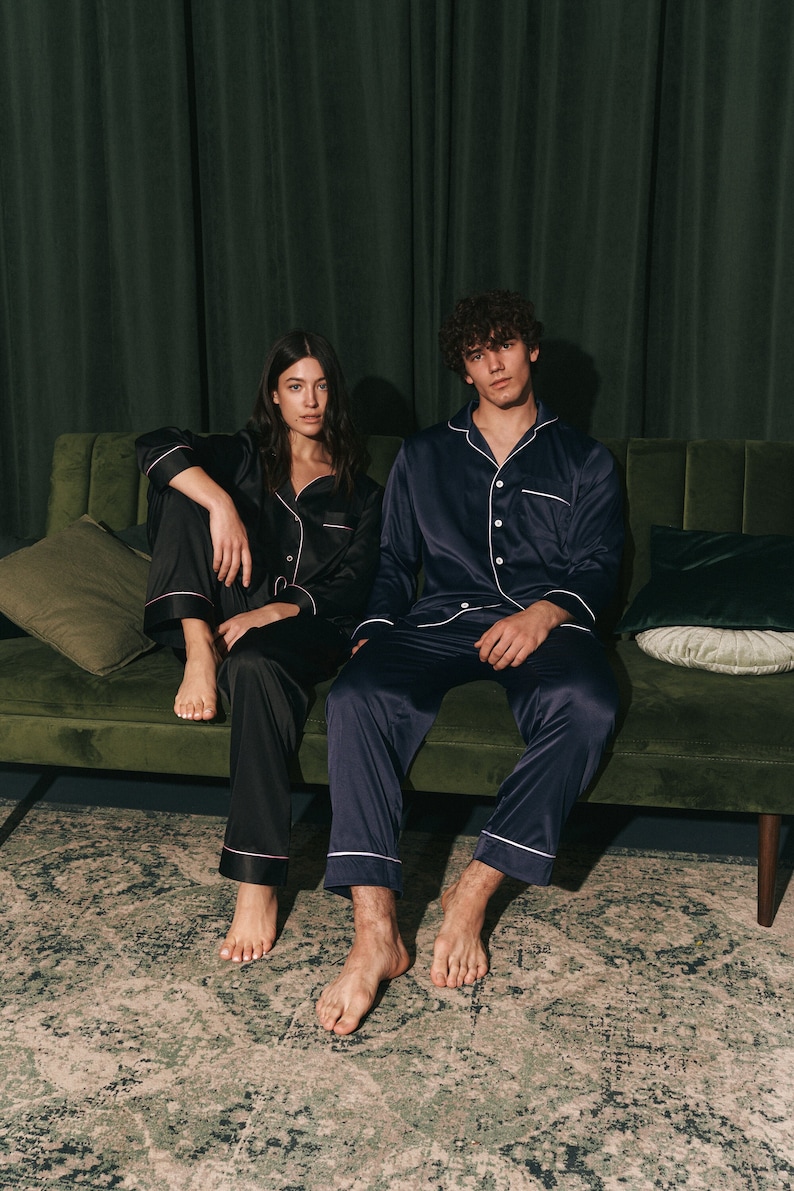 K&ouml;nnte beinhalten: Ein Paar tr&auml;gt passende schwarze und marineblaue Satin-Pyjamas und sitzt auf einem gr&uuml;nen Samtsofa. Die Frau tr&auml;gt ein schwarzes Pyjama-Oberteil mit wei&szlig;em Besatz und der Mann tr&auml;gt ein marineblaues Pyjama-Oberteil mit wei&szlig;em Besatz.