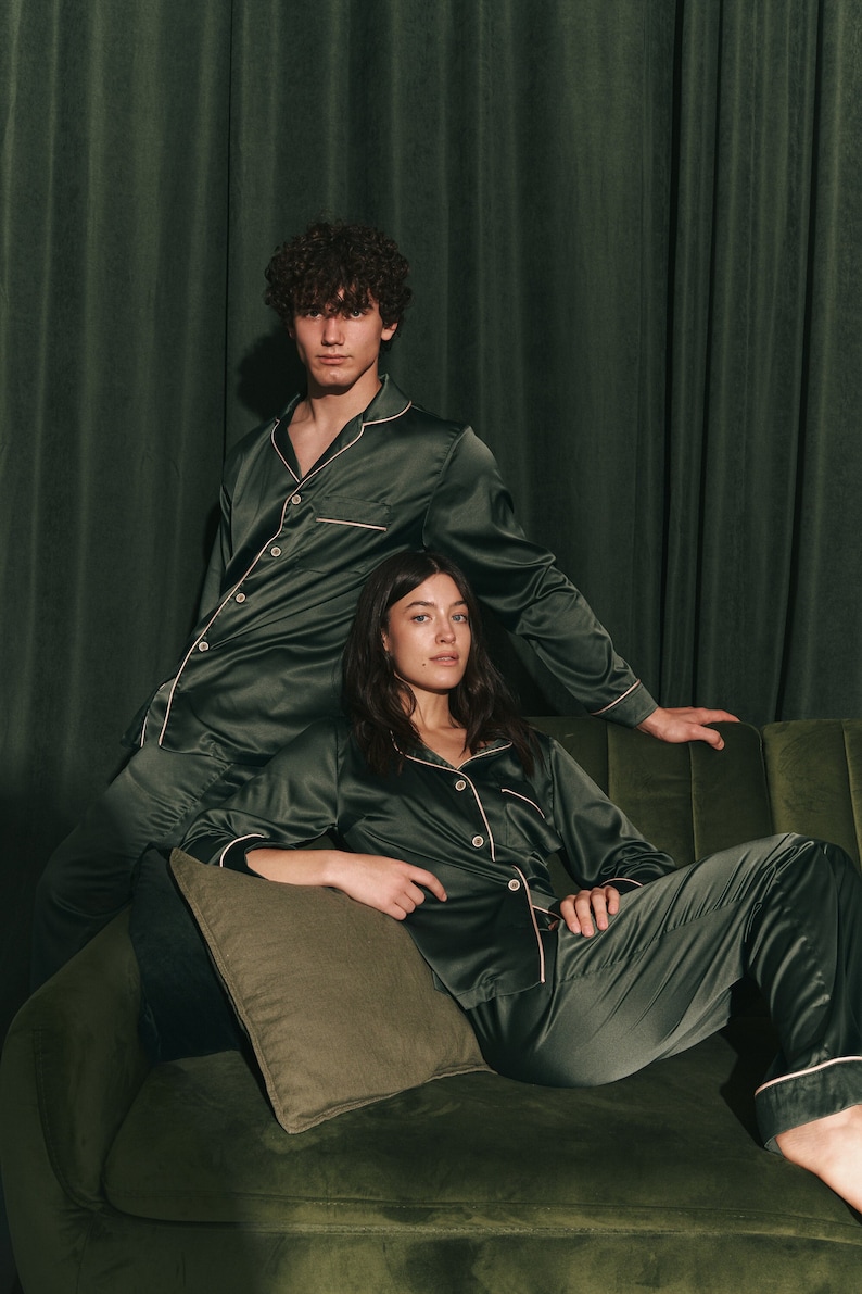 K&ouml;nnte beinhalten: Ein Paar tr&auml;gt passende dunkelgr&uuml;ne Satin-Pyjamas mit wei&szlig;en Paspeln. Sie sitzen auf einem gr&uuml;nen Samtsofa.