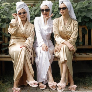 May include: Three women wearing white and gold bathrobes and pink bow slippers, with white towels wrapped around their heads, are sitting on a wooden bench. They are all wearing sunglasses and smiling.