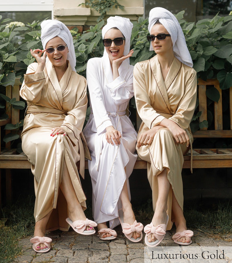 May include: Three women wearing white and gold bathrobes and pink bow slippers, with white towels wrapped around their heads, are sitting on a wooden bench. They are all wearing sunglasses and smiling.