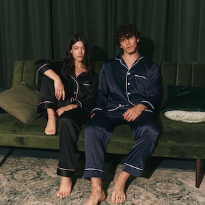 K&ouml;nnte beinhalten: Zwei Personen in passenden Pyjamas sitzen auf einem gr&uuml;nen Samtsofa. Die Frau tr&auml;gt einen schwarzen Pyjama mit rosa Paspeln und der Mann tr&auml;gt einen dunkelblauen Pyjama mit wei&szlig;en Paspeln. Beide schauen in die Kamera.