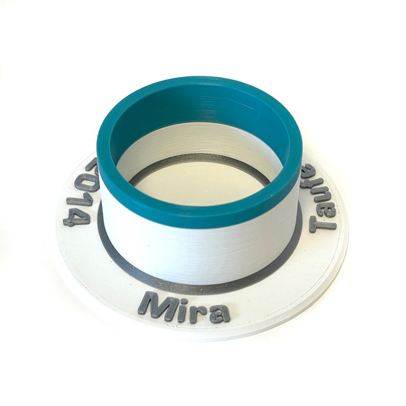 May include: White ceramic ring holder with a teal colored rim. The holder has the text "Mira" and "Tahel" on the sides and "2014" on the bottom.