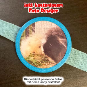 Könnte beinhalten: Ein kreisförmiger blauer Rahmen mit einem Foto eines cremefarbenen und eines schwarz-weißen Meerschweinchens, die Gras fressen. Der Rahmen ist an einem hellblauen Stoffband befestigt. Der Text oben lautet "inkl. kostenlosem Foto Desiger" und unten "Kinderleicht passende Fotos mit dem Handy erstellen!"