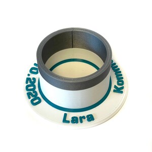 May include: A white and gray 3D printed cup with a teal rim and the text "Lara 2020 Kommun" printed on the side.