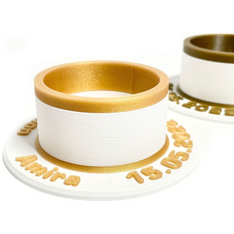 May include: White ring holder with a gold band and inscription "Amira 19.05.2021".