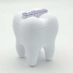 Könnte beinhalten: Ein weißer, zahnförmiger Behälter mit dem Wort "smile" in lavendelfarbenen Buchstaben auf der Oberseite. Der Behälter hat eine glatte, matte Oberfläche und ist so gestaltet, dass er einem Backenzahn ähnelt. Das Design ist schlicht und modern, passend für Zahn-Dekoration oder Aufbewahrung.