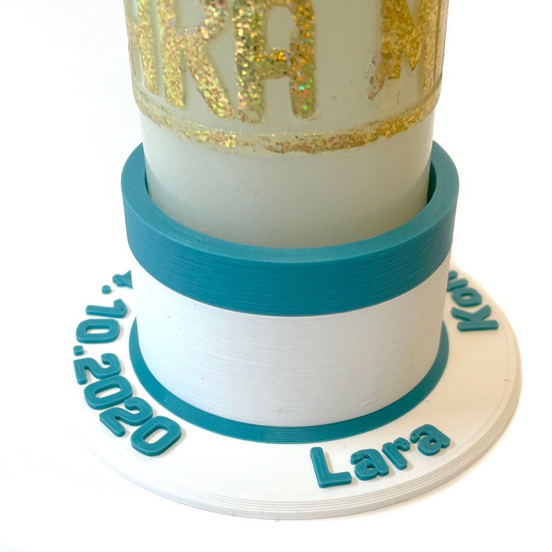 May include: White candle holder with a teal blue band and a white band with the text "Lara" and "10.2020" in teal blue.