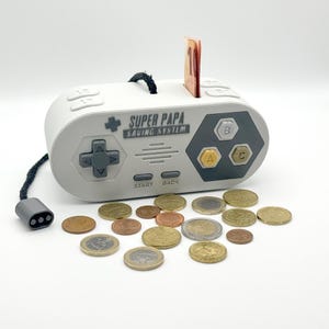 Könnte beinhalten: Ein weiß-graues Sparschwein in Form eines Videospiel-Controllers mit dem Text "SUPER PAPA SAVING SYSTEM". Einige Münzen und ein Geldschein sind sichtbar. Der Controller hat ein schwarzes Kabel und einen grauen Stecker.