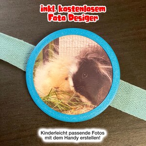 Könnte beinhalten: Ein kreisförmiger blauer Rahmen mit einem Foto eines cremefarbenen und eines schwarz-weißen Meerschweinchens, die Gras fressen. Der Rahmen ist an einem hellblauen Stoffband befestigt. Der Text oben lautet "inkl. kostenlosem Foto Desiger" und unten "Kinderleicht passende Fotos mit dem Handy erstellen!"