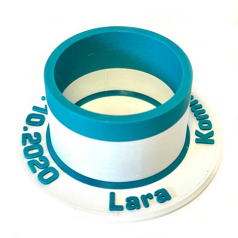 May include: A white and teal blue cylindrical container with a white base and a teal blue top. The container has the text "Lara 2020" and "Komi" printed on the base.