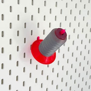Könnte beinhalten: Eine graue Garnrolle mit einem roten Plastikhalter, die an einer weißen Lochwand hängt.