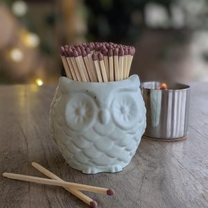 May include: A pale blue owl-shaped match holder filled with wooden matches. Two matches lie on the wooden surface. A small silver candle holder with a lit candle is to the right of the match holder.