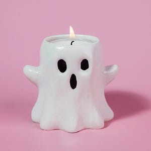 May include: A white ghost-shaped candle with black eyes and mouth, and a lit wick. The candle is set against a pink backdrop, perfect for Halloween. This handmade candle is a unique decorative item.
