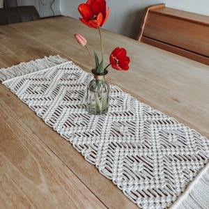 Könnte beinhalten: Ein weißer gehäkelter Tischläufer mit Fransen, der ein Diamantenmuster aufweist. Eine klare Glasvase mit goldener Oberseite hält rote Tulpen auf einem Holztisch.