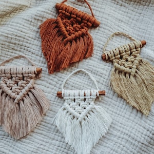 Könnte beinhalten: Vier kleine Makramee-Wandbehänge in Beige-, Braun- und Weißtönen. Jedes Stück hat ein gewebtes Design mit einem Holzdübel und einer Aufhängeschlaufe. Das Makramee hat einen Fransenboden. Die Aufhänger sind auf einer strukturierten, neutralfarbenen Oberfläche angeordnet.