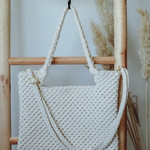 Peut inclure: Un sac cabas en crochet blanc avec une longue bandoulière suspendu à une échelle en bois. Le sac a un motif tissé et une fermeture en métal doré.