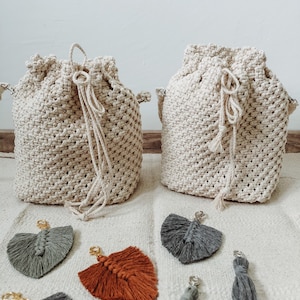 Peut inclure: Deux sacs à cordon en macramé beige avec un pompon sur chacun. Les sacs sont posés sur une surface blanche avec cinq porte-clés en macramé colorés.