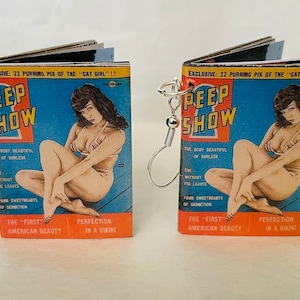 Może przedstawiać: Para kolczyków z miniaturami okładek książek z vintage'owym zdjęciem pin-up kobiety w bikini. Okładki książek są zatytułowane "Peep Show" i zawierają tekst "Exclusive: 22 Purring Pix of the "Cat Girl"!!".