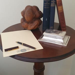 Peut inclure: Une table ronde en bois foncé présente une sculpture en bois, trois livres anciens, une boîte en argent, un stylo et un bloc-notes. Les livres sont intitulés "The Politics of Aristotle", "Atlas of Ancient Classical Geography" et "Stepping Heavenward". Le stylo et le bloc-notes sont sur la table.