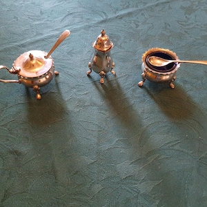 May include: A silver-toned condiment set consisting of a lidded mustard pot with a handle and spoon, a small perforated shaker, and an open salt cellar with a dark blue glass insert and a small spoon, all featuring three small feet.