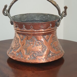 May include: A copper-coloured metal bucket with an ornate handle and decorative etched patterns. The bucket has a wide base and tapers upwards, with a detailed design around the body. The handle is a darker, aged metal.