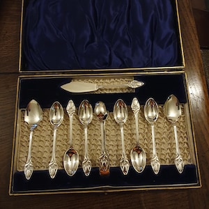 Puede incluir: Un juego de cucharas de plata vintage en una caja de presentación. El juego incluye cucharas y un cuchillo de mantequilla, todos con mangos ornamentados. La caja tiene un forro de satén azul y un marco dorado, creando una exhibición elegante.