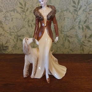 May include: A ceramic figurine depicting a woman in a brown coat with a fur collar and a cream dress, accompanied by a dog. The figurine is finely detailed and stands on a wooden surface.
