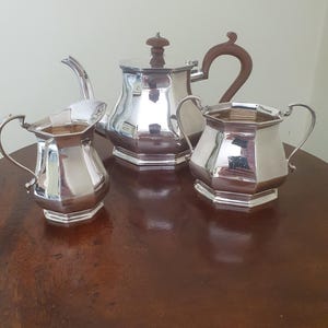 May include: A silver-plated tea set consisting of a teapot, a creamer, and a sugar bowl. Each piece features an octagonal body with a polished finish. The teapot includes a brown handle and a matching brown finial on the lid. The creamer and sugar bowl have curved handles and a matching octagonal base design.