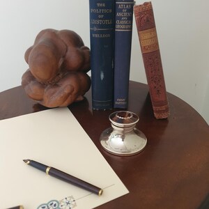 Könnte beinhalten: Ein Stillleben mit alten Büchern, einer Holzskulptur, einem silbernen Tintenfass und einem Füllfederhalter auf einem dunklen Holztisch. Die Bücher tragen die Titel "The Politics of Aristotle" und "Atlas of Ancient and Classical Geography."
