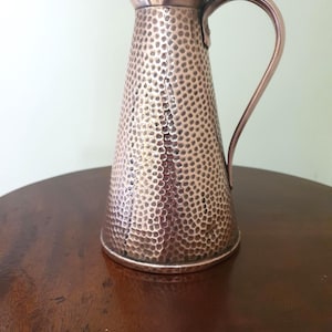 May include: A copper-coloured jug with a hammered texture. The jug has a curved handle and a spout for pouring. It is sitting on a dark brown wooden surface.