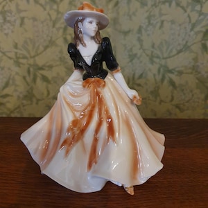 May include: A ceramic figurine of a woman in a long dress and a hat. The dress is cream with brown detailing, and the bodice is black. The hat is light grey with a brown brim. A decorative item, likely for display.