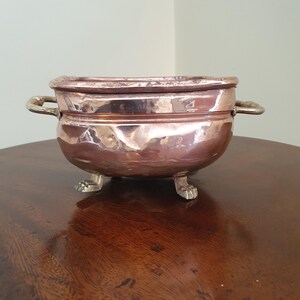 May include: A copper-coloured bowl with two handles and three small feet. The bowl has a rounded shape and a shiny surface. The handles and feet appear to be made of a different metal. The bowl is sitting on a dark wooden surface.