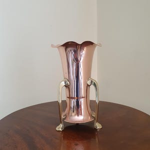 Könnte beinhalten: Eine kupferfarbene Vase mit geriffeltem Rand und zwei goldfarbenen Griffen. Die Vase hat eine reflektierende Oberfläche und steht auf einem dunklen Holztisch. Die Vase ist etwa 25 cm hoch.