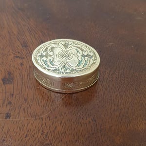 Puede incluir: Una pequeña caja ovalada de latón con un acabado dorado pulido. La tapa presenta un intrincado patrón floral y geométrico en relieve. El lateral de la caja también está decorado con un diseño grabado detallado y repetitivo en todo su contorno.