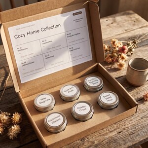May include: A cardboard box labeled "Cozy Home Collection" contains six small, round, silver-lidded tins of scented candles. The labels on the tins read "Wild Magnolia," "Dusk and Rose," "Sunday Morning Brew," "Cashmere Blanket," "Golden Crust," and "Sweet Violet Hour."