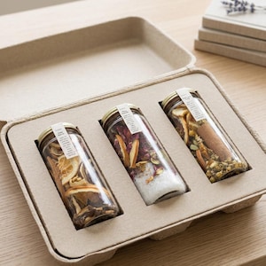 May include: Three glass jars filled with dried botanicals and spices, presented in a cardboard tray. The jars have gold lids and small labels. The tray is on a wooden surface, with a stack of books and lavender sprigs in the background.