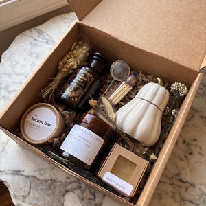 May include: A cardboard gift box containing a dark glass bottle labeled Autumn Brew, a round lotion bar, a jar of Exfoliating Cubes, a white ceramic pumpkin-shaped container, a small glass vial of matches, a tea infuser, and dried floral stems.