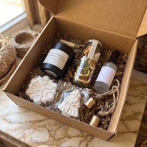 May include: A cardboard gift box filled with brown paper crinkle filler containing a black candle labeled Evergreen Enchantment, a glass jar of dried orange slices and greenery, a spray bottle, two white bath bombs, and two small gold cylinders.