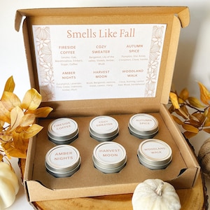May include: A cardboard gift box containing six small round metal tins with white labels. The lid interior features the text Smells Like Fall and lists scent profiles including Fireside Coffee, Cozy Sweater, Autumn Spice, Amber Nights, Harvest Moon, and Woodland Walk.